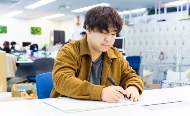 茶色のジャケットを着た男性スタッフが白いデスクでノートにアイデアを書き込む様子。オープンプランのオフィスで、モニターやロッカーが並ぶ現代的な職場環境