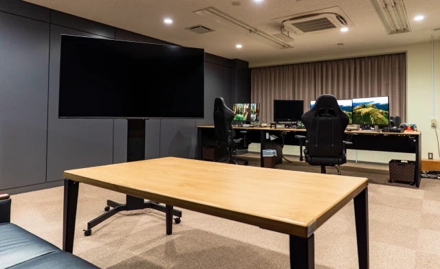 モダンで明るいオフィススペース。中央に大型ディスプレイ、手前に木製の会議テーブル、背景に複数モニターを備えたワークステーションが配置された快適な職場環境