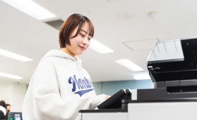 ショートボブの女性スタッフが大型のオフィスプリンターを操作する様子。明るく清潔感のあるオフィス空間で、快適に業務に取り組む職場の雰囲気