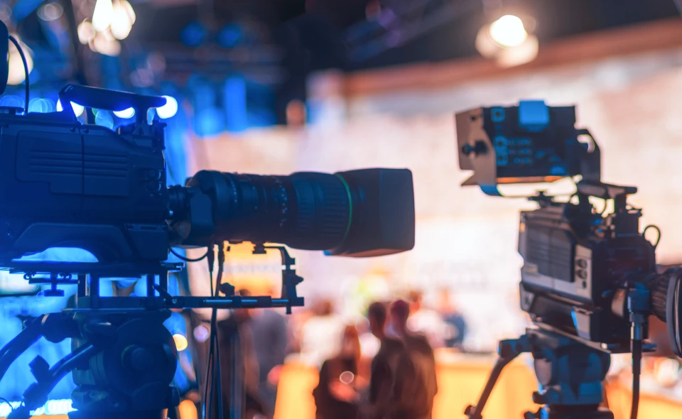 イベント会場で、青とオレンジの照明を背景に三脚に設置された2台のプロ用ビデオカメラ