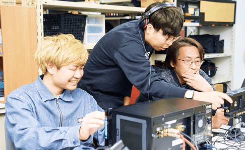 3人の男性スタッフが電子機器を囲んで真剣に打ち合わせや作業を行う様子。技術的な課題に協力して取り組むチームワークと専門性が感じられる職場環境