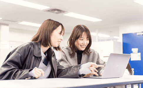 2人の女性スタッフがラップトップを囲んで笑顔で協力しながら作業する様子。明るく開放的なオフィス環境で、チームワークを大切にする職場の雰囲気