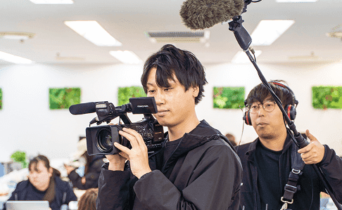 プロフェッショナルなビデオカメラを操作するカメラマンと、ブームマイクを持つ音声スタッフが協力して映像制作に取り組む様子。現場でのチームワークと専門性が感じられる制作現場